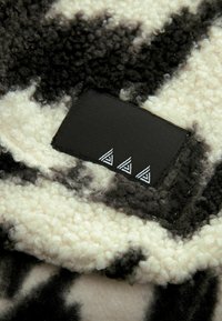 Blød fleece-tæppe i sort, hvid og grå med en struktureret, høj-plysoverflade. Har et sort mærke med tre hvide trekantgrafikker.