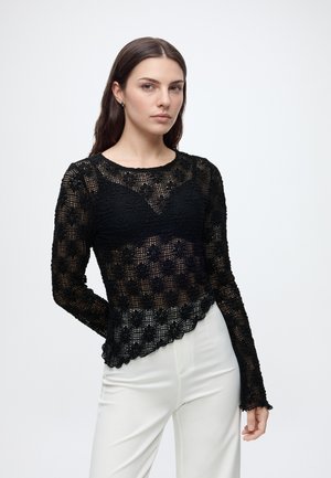 Femme aux longs cheveux foncés portant un haut noir en dentelle florale à manches longues et un pantalon blanc, debout devant un fond clair uni.