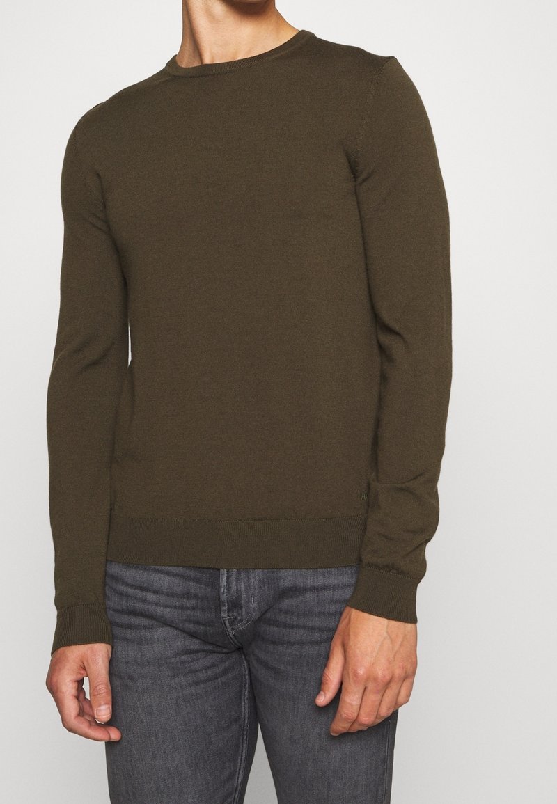 Dunkelgrüner Rundhalsausschnitt-Pullover aus weichem Strickmaterial. Mit langen Ärmeln und geripptem Saum sowie Bündchen. Getragen mit grauen Jeans.