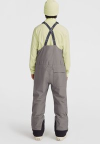 Grijze snowboard bibbroek met verstelbare zwarte banden, uitgerust met zakken en een losse pasvorm, gecombineerd met een lichtgroene langekleding en een pet.