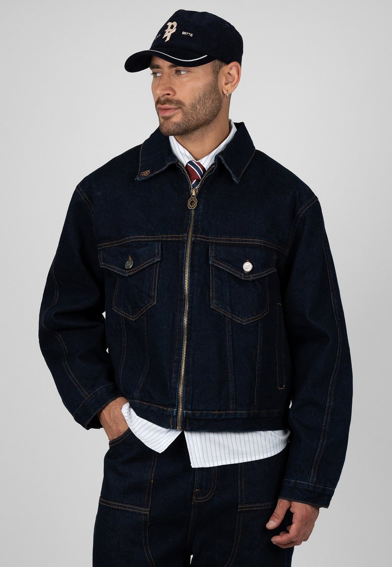 Homme portant une veste en jean foncé, un jean assorti, une chemise blanche rayée, une cravate rouge rayée et une casquette noire, regardant sur le côté avec une main dans la poche.