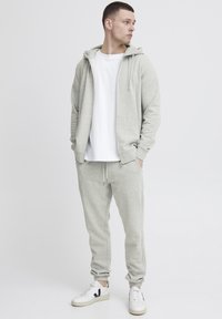 Hellgrauer Hoodie mit Frontreißverschluss und Kordelzügen, kombiniert mit passenden Jogginghosen. Darunter ein weißes T-Shirt und weiße Sneakers.