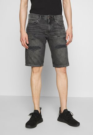 Person trägt schwarze, abgenutzte Jeans-Shorts, schwarze Sneakers und ein schwarzes Hemd, steht vor einer schlichten grauen Wand und einem grauen Boden.