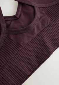 Bordo spalvos sportinė liemenėlė su tekstūruotomis detalėmis, pagaminta iš minkšto audinių mišinio ir plačiu apačios dirželiu. Viršuje matomos dirželių kontūrai.