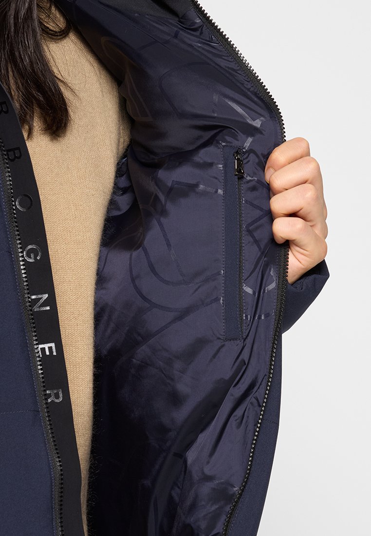 Person, der eine navyfarbene Jacke aufhält, die eine Innentasche mit Reißverschluss und schwarze Markenverzierungen entlang des Reißverschlusses zeigt, darunter einen beigen Pullover.