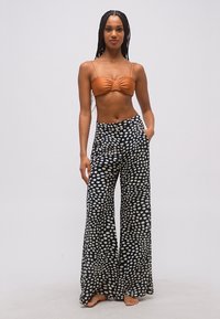 Moteris su ilgais kaspinais, vilkinti oranžinį trumpą viršutinį drabužį ir juodus, platėjančius, aukšto liemens kelnes su baltais taškeliais, stovinti basomis.