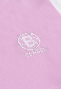 Camisa de tela rosa con un logo bordado redondo en blanco que presenta una "B" estilizada y la palabra "BESSEL" debajo. Textura suave.