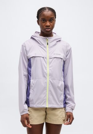 Žena v svetlofialovej kapucňovej bunde Columbia so zeleným zipsom a béžovými krátkymi nohavicami stojaca pred bielym pozadím.