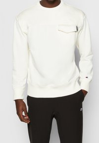 Felpa color crema con scollo rotondo, tasca frontale, polsini e orlo a coste; abbinata a pantaloni scuri con un accento logo.