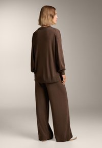 Barna kötött pulóver és bő szabású nadrág szett, puha textúra, laza szabás, hosszú ujjak és mandzsetták. Megjegyzés: cipők nincsenek hangsúlyosan kiemelve.