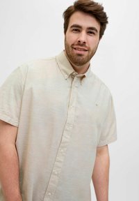 Chemise à manches courtes, boutonnée, en beige clair, avec une texture subtile et une poche ornée d'un petit logo.