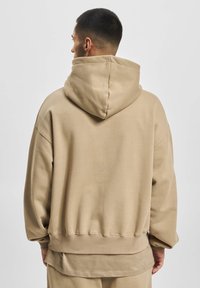 Haut de survêtement oversize beige avec une grande capuche, des épaules tombantes, des poignets côtelés et une finition douce et texturée. Conçu pour le confort et la chaleur.
