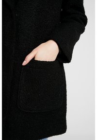 Manteau texturé noir avec une grande poche plaquée. Le matériau semble duveteux, avec une coupe ample et des manches larges pour plus de confort.