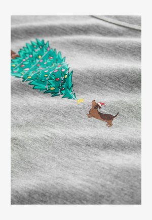 Pelēkā auduma virsmā attēlots zaļš Ziemassvētku koks, kas izrotāts ar krāsainiem rotājumiem, un brūns suns, kas valkā Ziemassvētku vecīša cepuri, sasniedz zvaigzni.