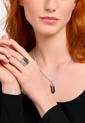 Rothaarige Frau mit schwarzem Oberteil zeigt einen silbernen Ring und passenden Anhänger mit Tigeraugen-Steinen.