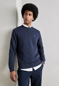 Marine crewneck sweatshirt in een katoenmix, met ribgebreide cuffs en zoom. Bevat een klein vierkant logo op de borst. Draagt over een wit overhemd met kraag.