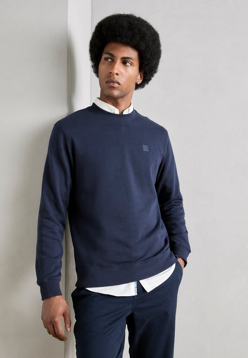 Marine crewneck sweatshirt in een katoenmix, met ribgebreide cuffs en zoom. Bevat een klein vierkant logo op de borst. Draagt over een wit overhemd met kraag.