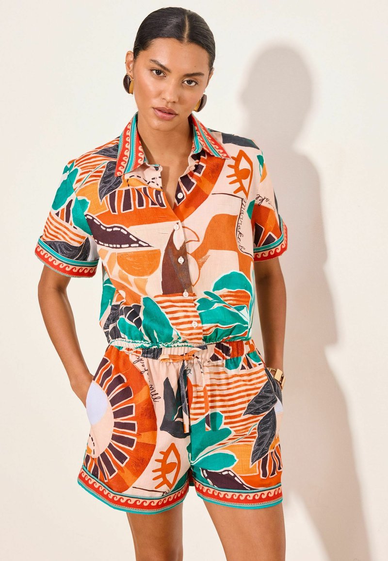 Woman wearing colorful patterned short-sleeve button-up shirt and matching shorts with orange, green, and black abstract designs, hands in pockets.
