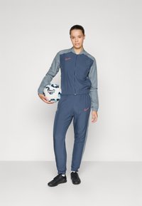 Nike atletisk træningsdragt i navy og grå, med en zip-up jakke, tapered bukser og en fodbold med et geometrisk mønster.