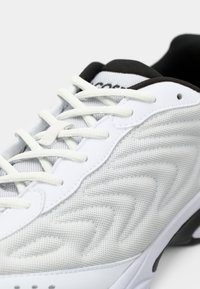 Chaussure de sport blanche avec texture en mesh, lacets blancs, doublure intérieure noire et motifs ondulés sur le côté en gros plan.