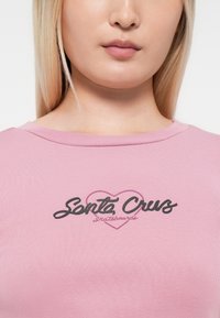 Rožinė ribota marškinė su juodu ir rožiniu širdies kontūru, kuriame per krūtinę užrašyta "Santa Cruz Skateboards" šriftu, kuris imituoja kursyvą.