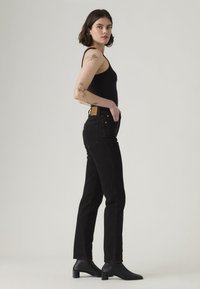 Black denim jeans with a high waist, featuring a brown leather patch on the back, styled with a fitted black tank top and black heeled boots.