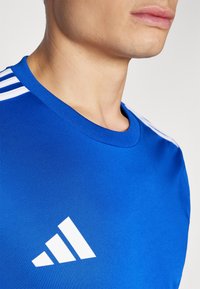 Blue athletic shirt with a round neck and white stripes on shoulders. Features a white triangular logo on the left side. Smooth fabric texture.