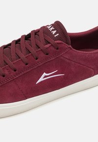 Μπορντό σουέτ αθλητικά παπούτσια με λευκό λογότυπο, υφασμάτινη γλώσσα με υφή, επίπεδες κορδόνες και λευκή ελαστική σόλα. Απλός, χαμηλού προφίλ σχεδιασμός.