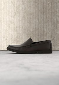 Čierny kožený slip-on mokasín s nízkym podpätkom, zaoblenou špičkou a minimálnym prešívaním, umiestnený na textúrovanom pozadí s vertikálnymi pruhmi.