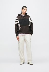 Fiatal férfi fonott hajjal, fekete-fehér Adidas kapucnis pulóverben, fehér Adidas nadrágban és fehér sportcipőben, egyszínű háttér előtt.