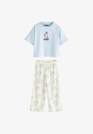 Barn med langt, krøllet hår står barfodet iført en lyseblå Mummitrold T-shirt og hvide pyjamasbukser med fårmønster i et blødt oplyst rum.
