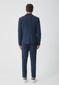 Ναυτικό κοστούμι με δομημένο σακάκι και ραμμένο παντελόνι, συνδυασμένο με λευκά αθλητικά παπούτσια με ναυτικές λεπτομέρειες. Λεπτό ύφασμα και καθαρές γραμμές.