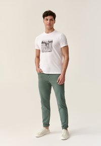 Camiseta blanca con un gráfico de bicicleta y el texto "LA BICICLETA EN MIS GENES", combinada con pantalones verdes y zapatillas de color claro.