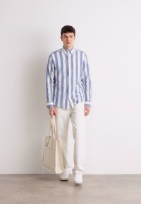 Gestreepte blauwe en witte overhemd, lichte broek en witte sneakers. Draagt een beige tote bag. Eenvoudig ontwerp, casual uitstraling.