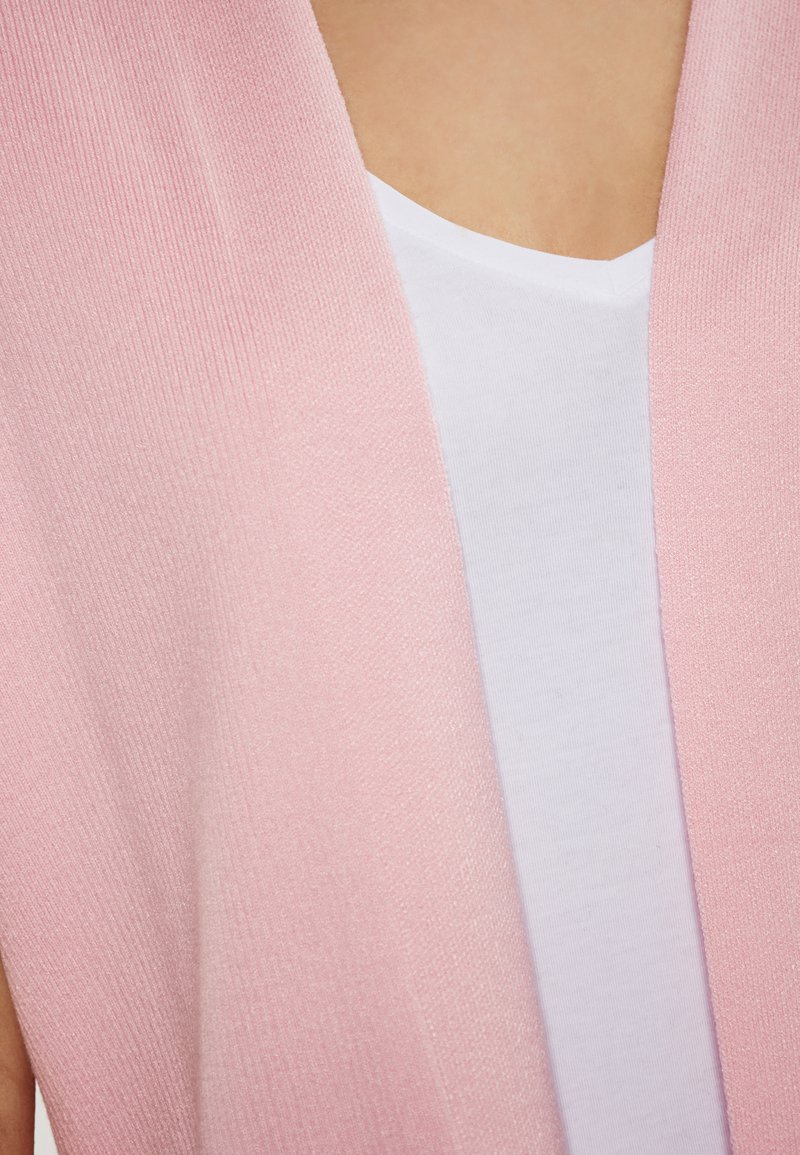 Hells rosefarbener offener Cardigan aus strukturiertem Stoff, der sanft über einem weißen V-Ausschnitt-T-Shirt fällt. Auffälliger Farbkontrast und weiche Textur.