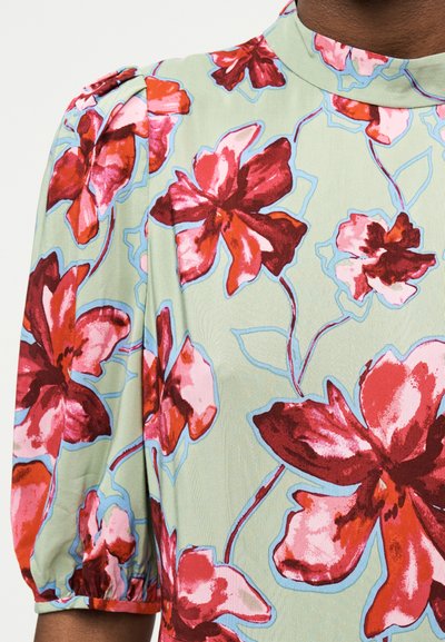Κοντινό πλάνο κοντομάνικου ενδύματος με κόκκινο και ροζ ανθικό σχέδιο σε ανοιχτοπράσινο ύφασμα.