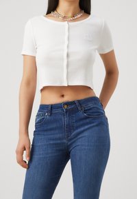 Haut court en ribbed blanc à manches courtes avec des détails de boutons et un petit logo brodé, associé à un jean en denim bleu ajusté.