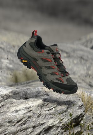 Chaussure de randonnée vert foncé et rouge avec semelle Vibram robuste suspendue au-dessus d’un terrain extérieur rocheux avec de petites plantes.
