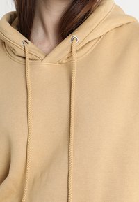 Sudadera con capucha beige, con cordón y ojales plateados. Fabricada en un tejido suave con una textura lisa.