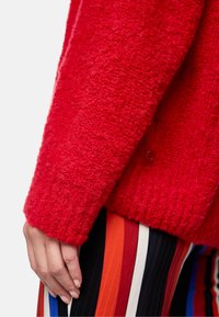 Roter, strukturierter Strickpullover mit lockerer Passform und gerippten Bündchen. Verfügt über einen Knopfdetail am Ärmel. Kombiniert mit mehrfarbigen gestreiften Hosen.