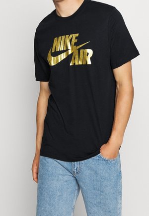 T-shirt en coton noir avec un grand logo "NIKE AIR" doré en impression lisse et audacieuse. Coupe standard avec manches courtes, porté avec un jean bleu clair.