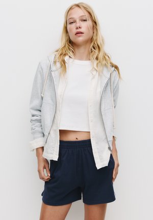 Felpa grigia con cerniera, crop top bianco e pantaloni corti elasticizzati blu scuro. Tessuto leggero, vestibilità rilassata, con dettagli del cordoncino sulla felpa.
