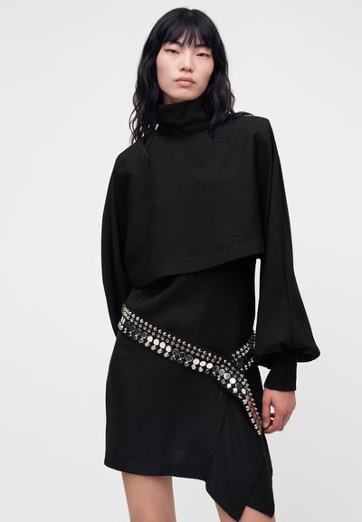 Modèle aux longs cheveux foncés portant un haut drapé noir et une jupe, soulignés par une ceinture diagonale ornée de clous et de disques argentés, sur un fond uni.