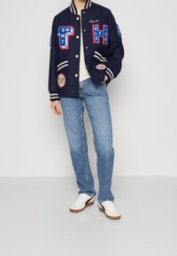 Chaqueta varsity azul marino con acentos en blanco y rojo, detalles de bordado, puños y cuello de canalé. Llevada sobre una camisa color crema y jeans.