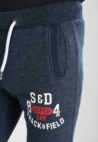 Donkerblauwe sweatpants van een gestructureerde stof, met een elastische tailleband, witte trekkoord, zijzakken en een geprint logo in wit en rood.