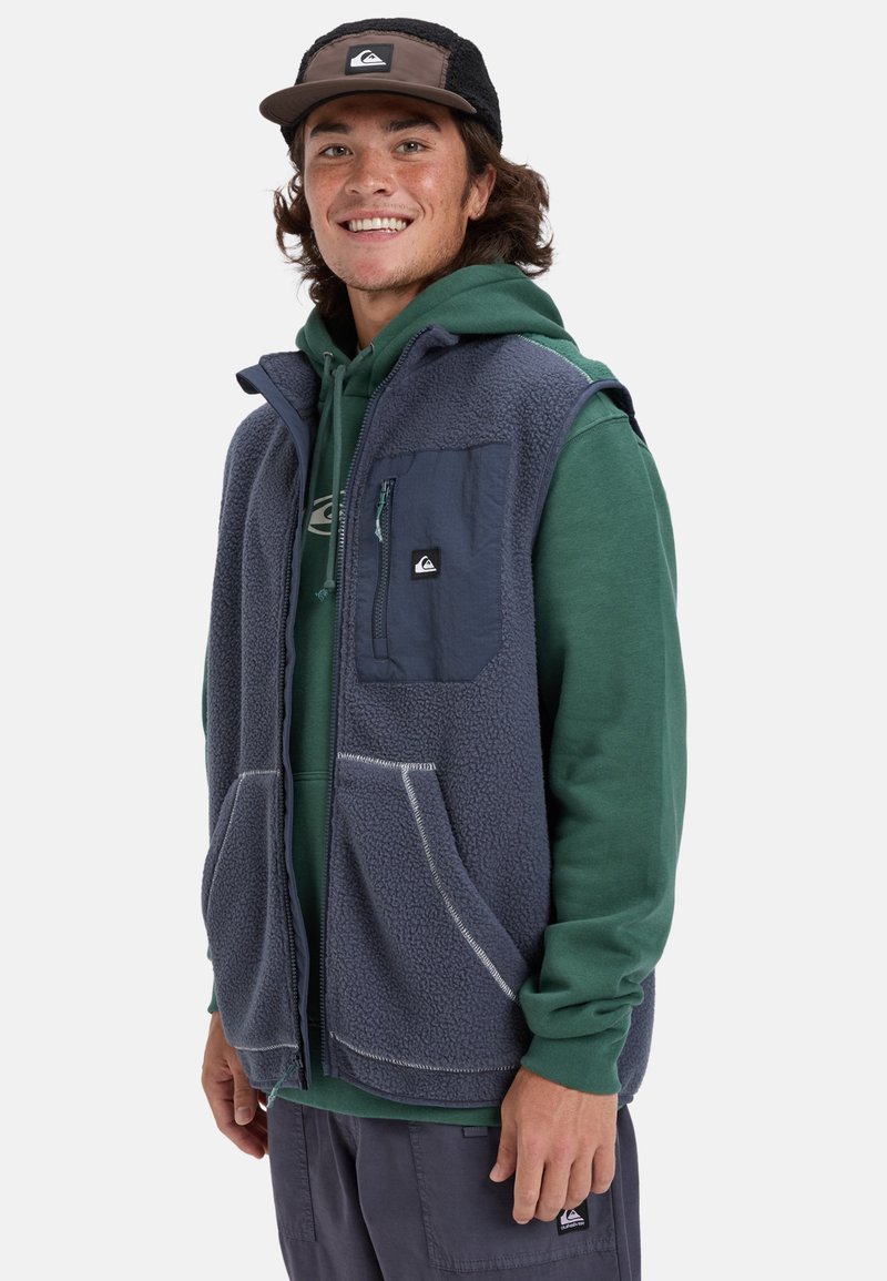 Grå fleeceväst med en blå ficka och vita sömnadsdetaljer, lagd över en grön hoodie, med en logo på bröstfickan.