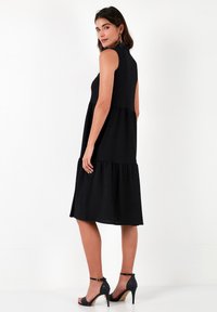 Black sleeveless dress with a tiered design, a high neckline, and a soft texture. Worn with black high-heeled sandals, showing a clean silhouette.