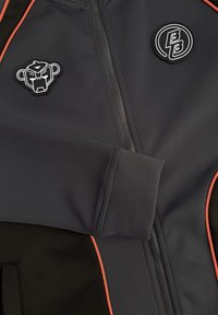 Donkergrijze zip-up jas met oranje piping, geribde manchetten en twee zwart-witte patches met een gestileerd berenhoofd en elkaar overlappende letters.