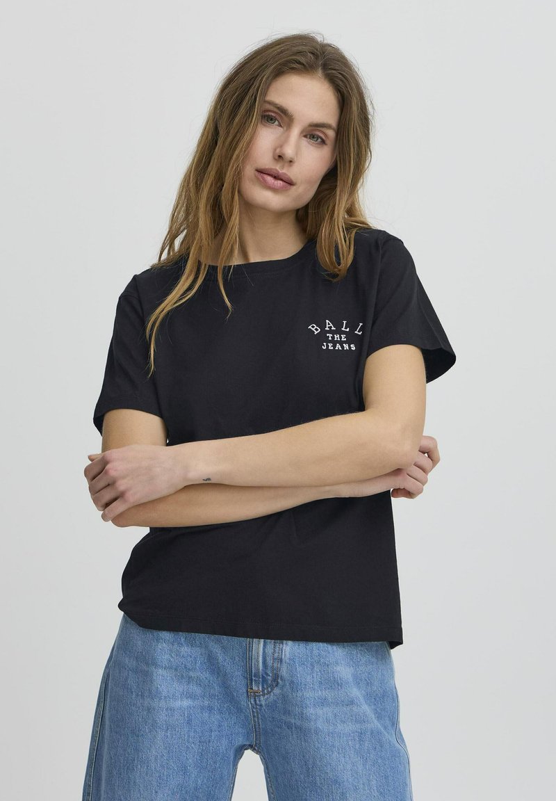 Tricou negru din bumbac cu mâneci scurte, având text alb care spune "BALL THE JEANS" pe pieptul stâng. Poartă o pereche de blugi de denim de culoare bleu deschis.