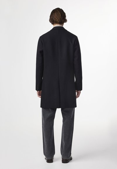 Musta villatakki, jossa on räätälöity leikkaus ja lovettu kaulus. Ominaisuuksina yksittäinen halkio ja nappikiinnityksellä varustetut hihat. Pukeutunut harmaisiin housuihin ja tummiin kenkiin.